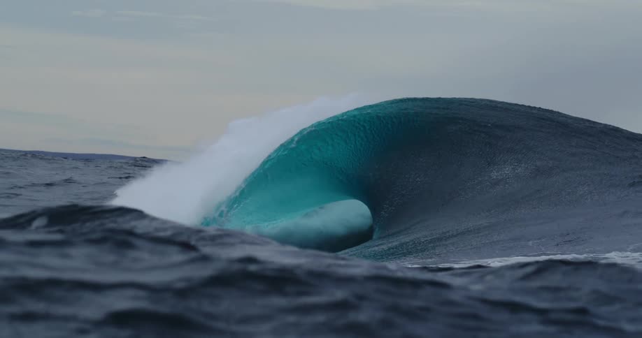 A huge blue wave curls and splashes as it rolls on rough water under a cloudy sky