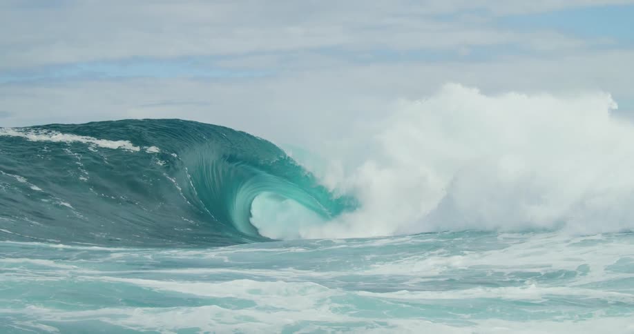 Water splashes as a huge blue wave curls as it rolls in under a cloudy sky