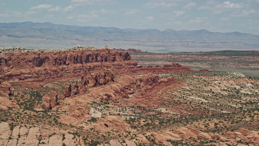 Aerial view of Klondike Bluffs and Marching Men rock formations in Arches National Park Utah with canyons stretching across the land and mountains in the distance under a cloudy sky during a summer day