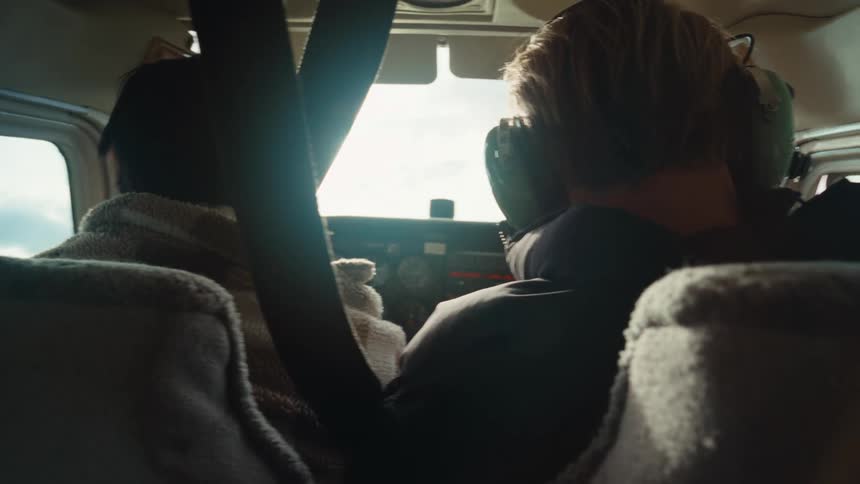 Two people sit in the cockpit of small plane flying over treetops