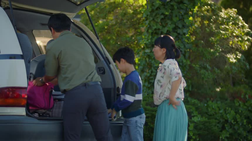 Man taking the suitcase handed to him by his son and wife and placing it in the cars trunk