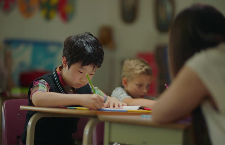 Two boys and a girl indoors sitting at a desk in a classroom coloring with pencils and one of the boys stopping to stare with a puzzled facial expression at the girl