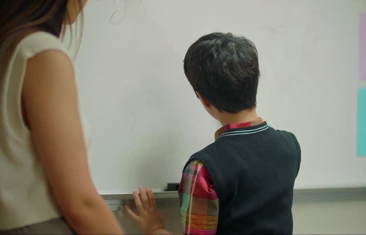 A child writes on the classroom board while the teacher watches