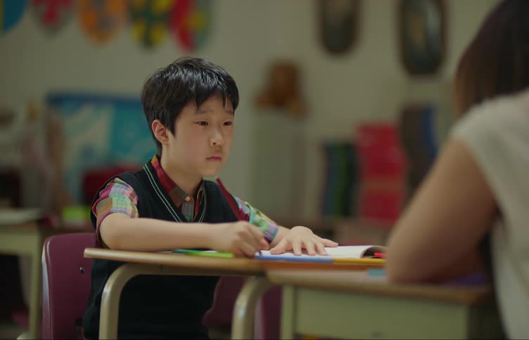 A student sitting at the desk drawing with their teacher in front of them having a conversation