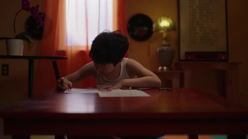 Boy in a tank top sitting at a table and drawing on a sheet of paper