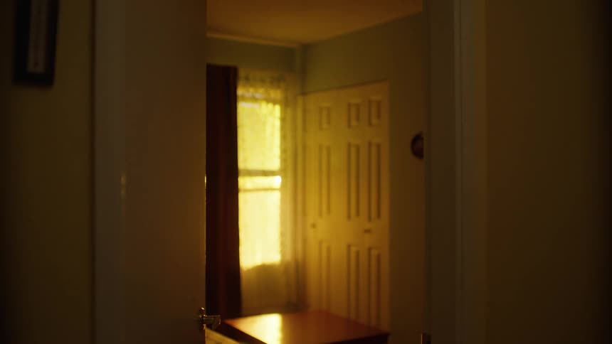 A young boy exits the living room leaving the door ajar the sunlight reflects off the window which is adorned with a white translucent curtain