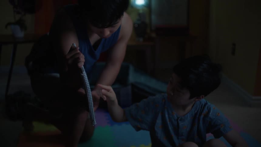 Child sitting and watching television when their older brother arrives and asks them a question from the notebook the child points their finger and responds promptly