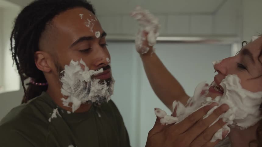 A young man and woman in a bathroom apply shaving cream all over each others faces