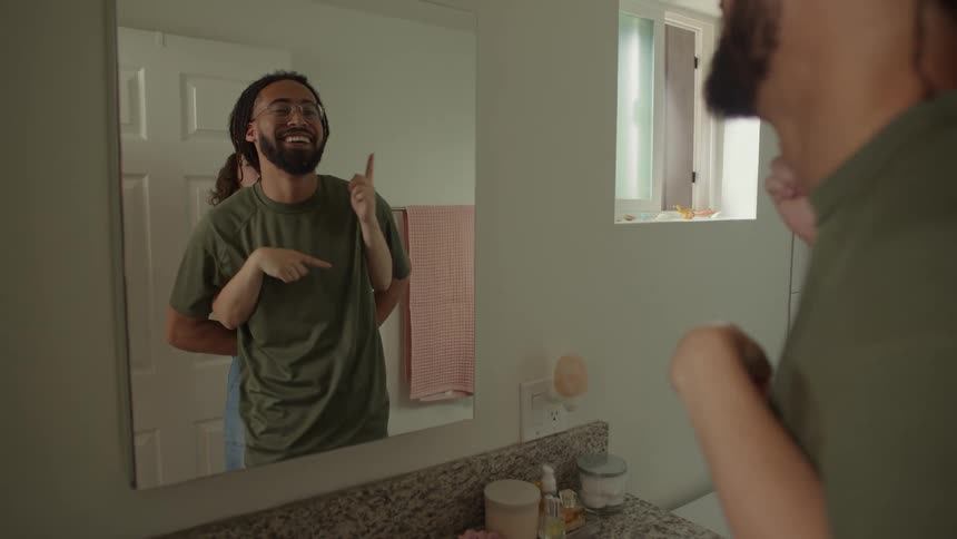 Man with dreadlocks glasses and beard and woman with curly hair look in the mirror mimicking each other in the bathroom