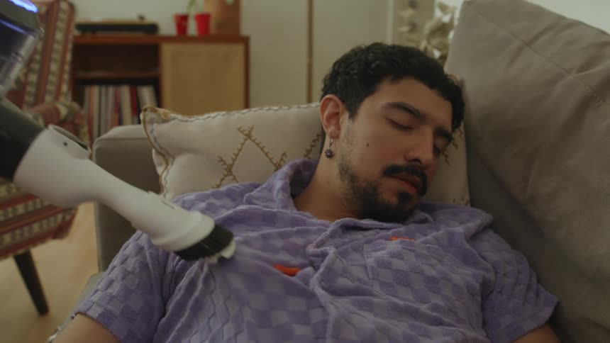 Man sleeping on a couch while a vacuum cleaner is sucking up food from his clothes