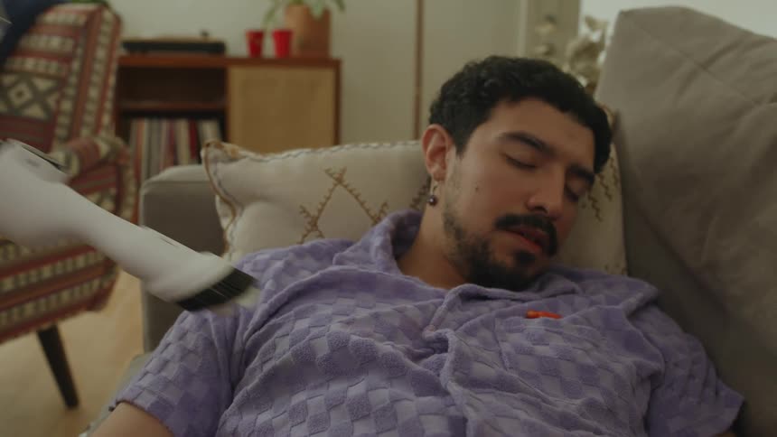 A man sleeping on the couch while a vacuum cleaner is sucking up an orange object on top of his clothing
