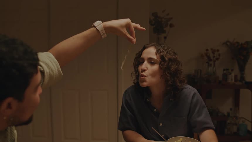 Friends sitting at a table with a man holding a strand of pasta and putting it into a womans mouth