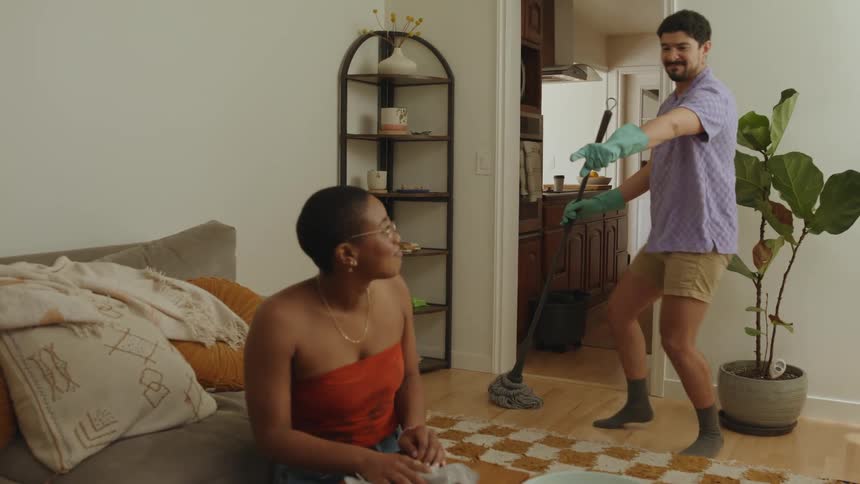 A man walks in the room mopping as a woman cleans a coffee table full of food