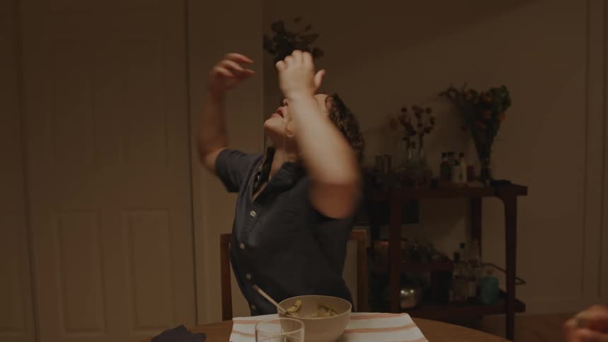 Friends sitting at a dinner table with food and one of them picking up a strand of pasta and playfully putting it on their face while they talk and smile