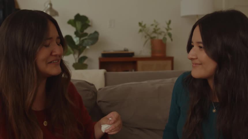 Two friends sitting on the couch smiling and chatting with each other while one of them passes a tissue to the other