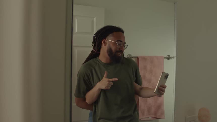 A woman hiding behind a man with a beard glasses dreadlocks and a green tshirt pulls a prank with a smartphone in the bathroom