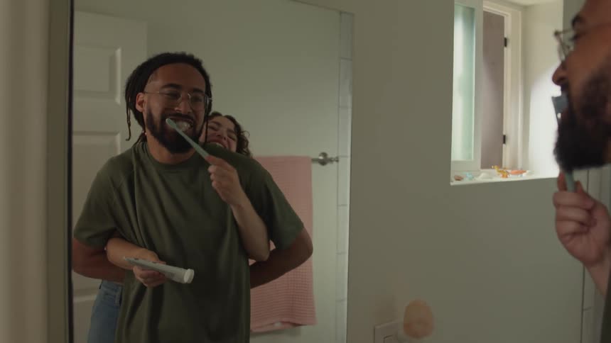 Man and woman stand near bathroom mirror brushing teeth laughing