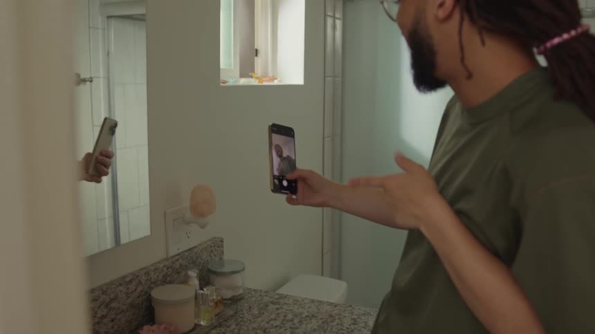 A man is standing in his bathroom holding his phone and smiling and making hand gestures as his girlfriend stands behind him