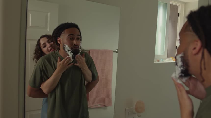 Woman applies shaving cream to mans face standing in front of mirror