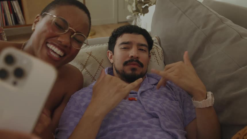 A man is lying on the couch as a woman sitting next to his holds up her phone and they laugh as he makes hand gestures