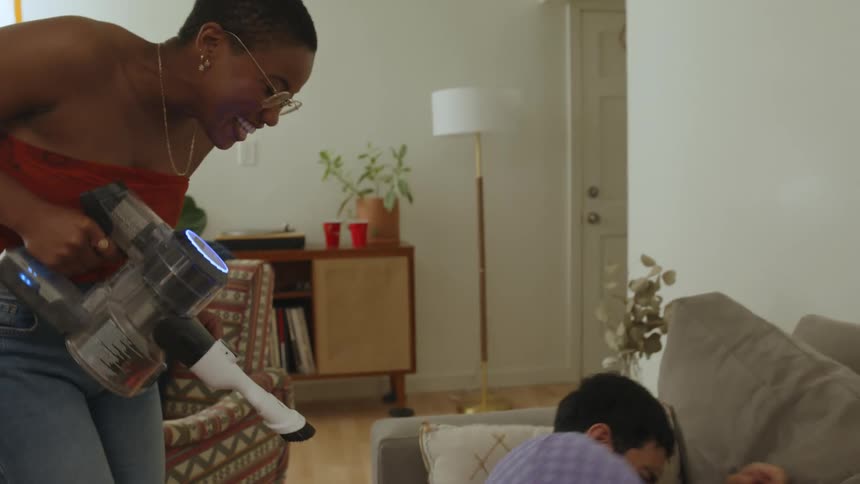 Woman cleaning with a vacuum playfully disturbs a man napping in the couch