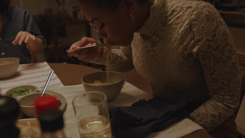 Two friends sitting at a table with one of them watching the other eat pasta and both of them smiling a lot