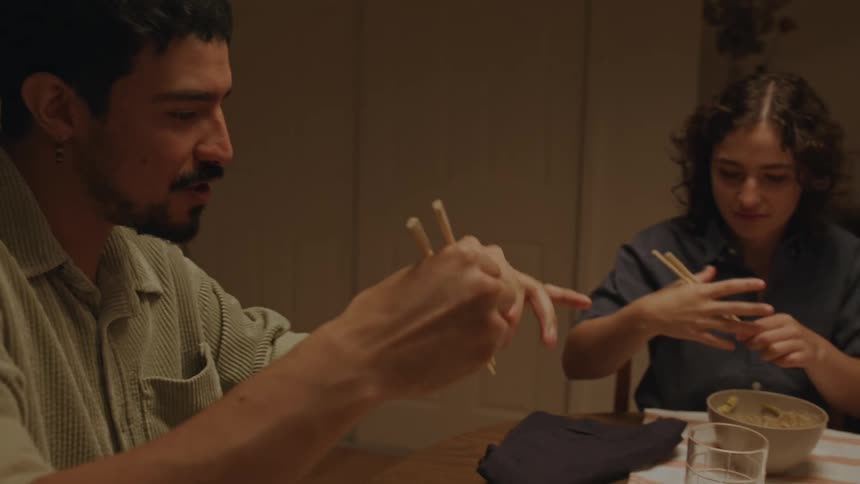 A man and a woman sitting at a dinner table try to teach a teenager to eat with chopsticks