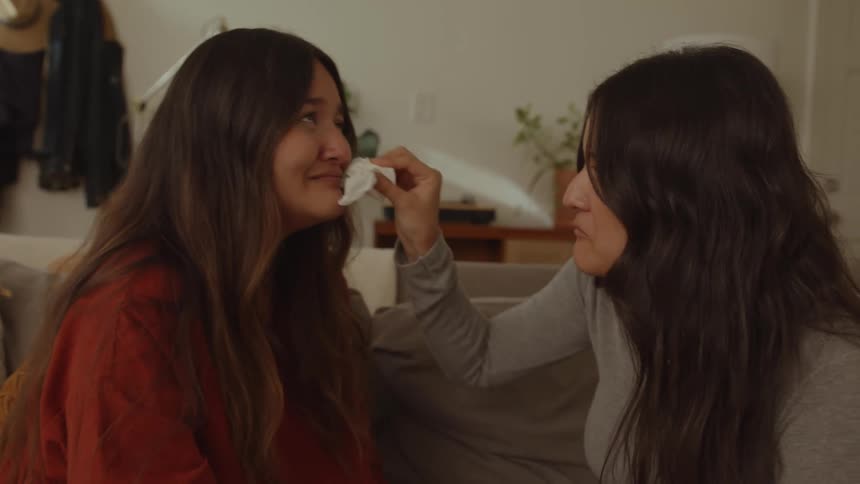 One woman helps the other woman wipe her tears as they sit laughing on couch