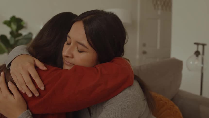 A sad woman cuddles her friend who is sitting on the couch in the living room