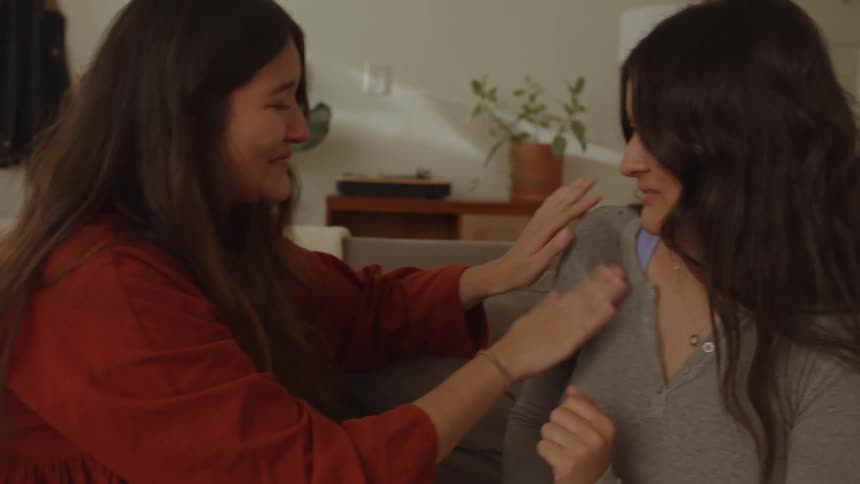 Two friends sitting on the couch with one of them crying emotional and happy tears wiping the wet shirt of her friend