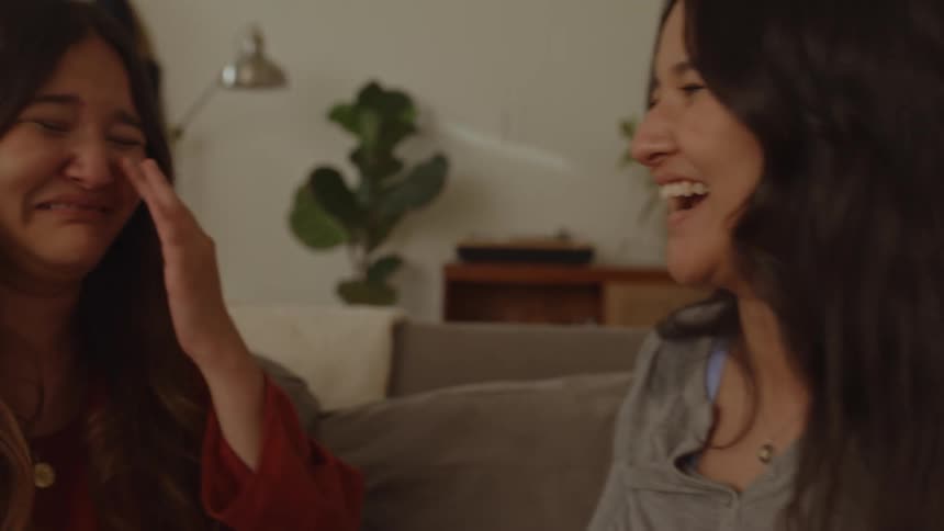 Two friends sitting on the couch smiling and emotional with one of them using her shirt to wipe away the tears of the other