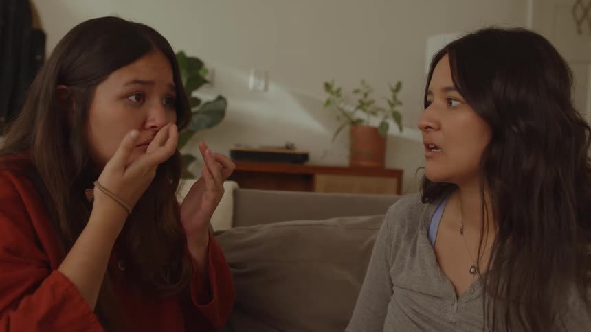 Two women sit on couch indoors one wipes tears from eyes