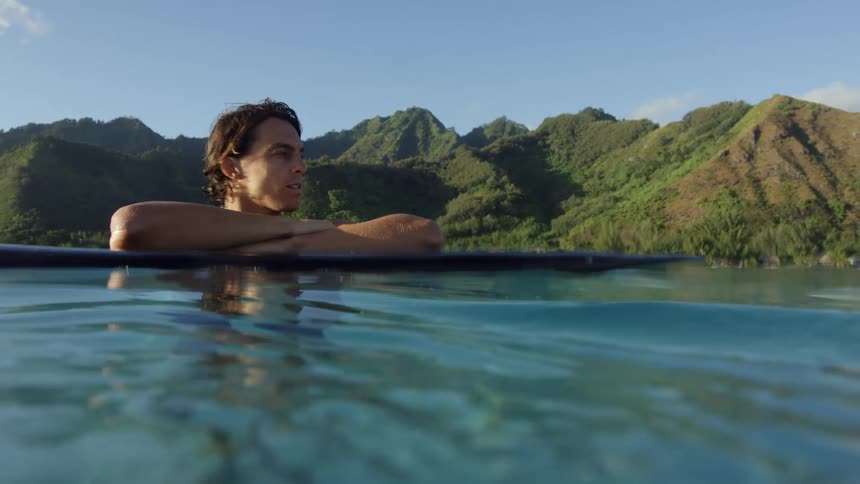 A man inside the sea water hanging his hands on his surf board while looking to his left with grass covered hills on shore