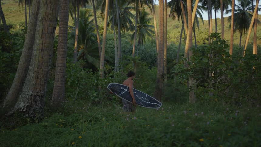 Shirtless man walking through the forest at midday while carrying a surfboard