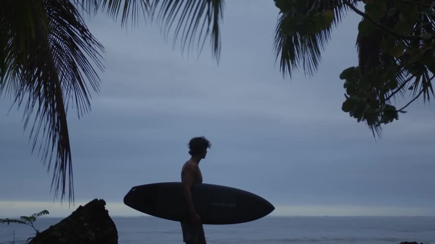 Man walking on the beach holding the surfboard and observing the sea