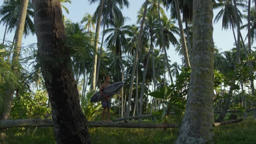 Man holding his surfboard atop a tree stump in a tropical environment