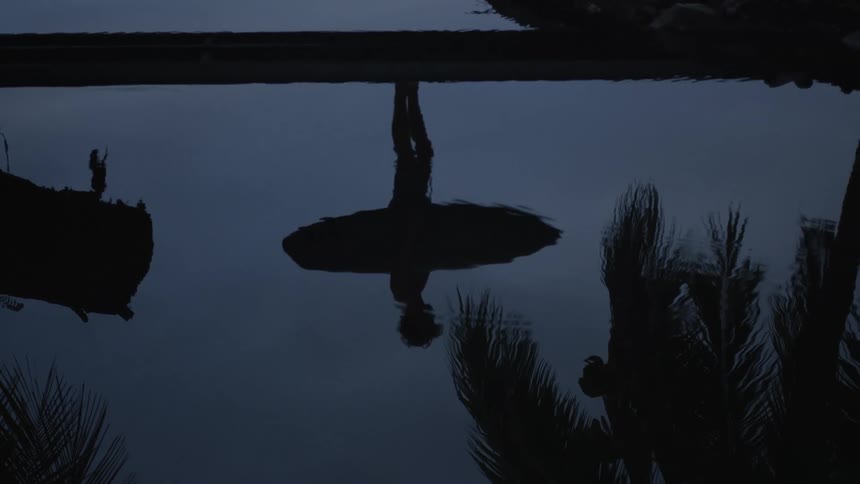 A man reflection is seen through the water holding a surf board and standing