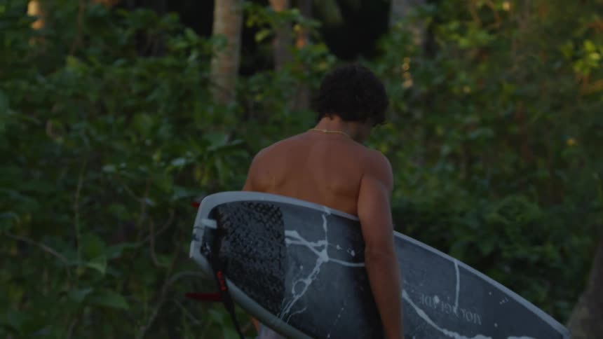 A young man walks through the woods with a black surfboard