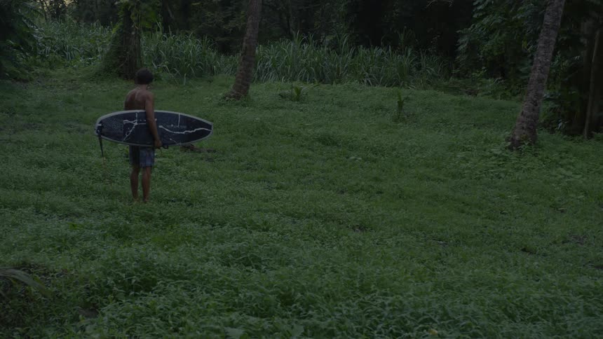 A man in a natural environment with grass and trees holding his surfboard