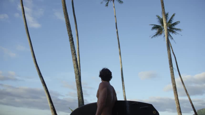 A man is standing near palm trees holding his surfboard