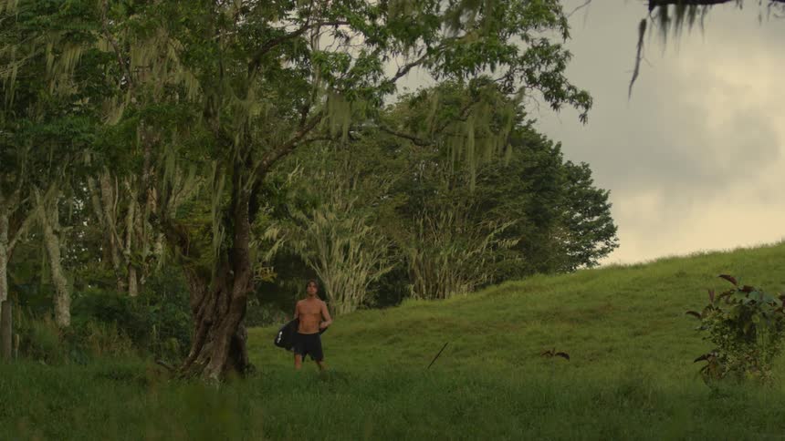 Young shirtless man carries a surfboard through the hills