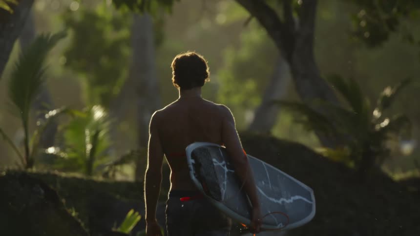 A man walks with a surfboard in the woods