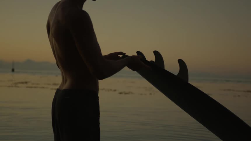 Man waxing his surfboards fins in the sea at dawn
