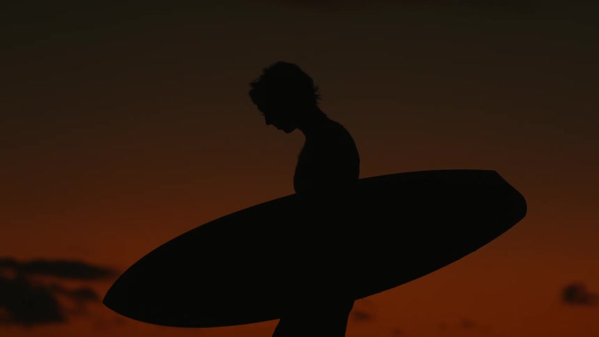 Man holding his surfboard and walking on the beach with a reddish sky