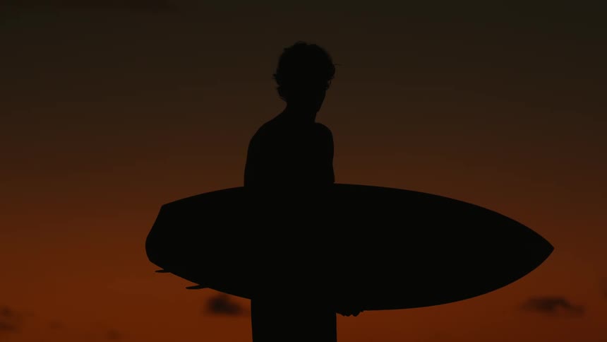 Silhouette of a man walking with his surfboard against an orange sky
