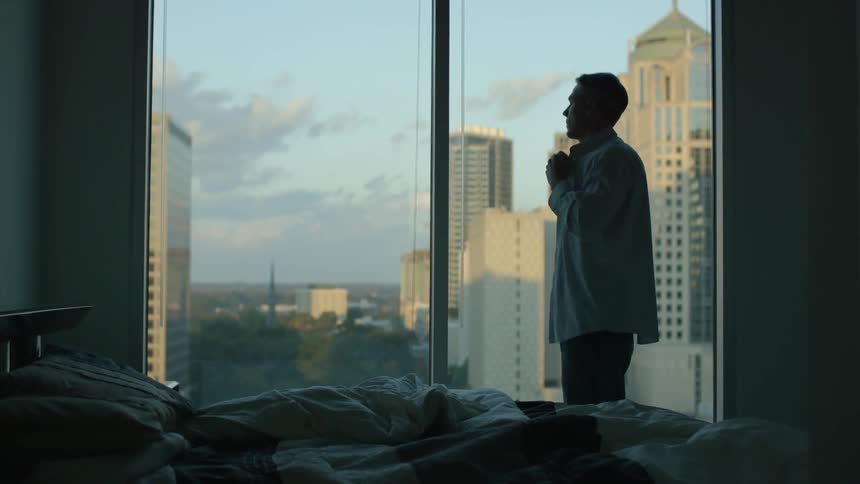 Man buttoning up his shirt in front of a large window in a big city