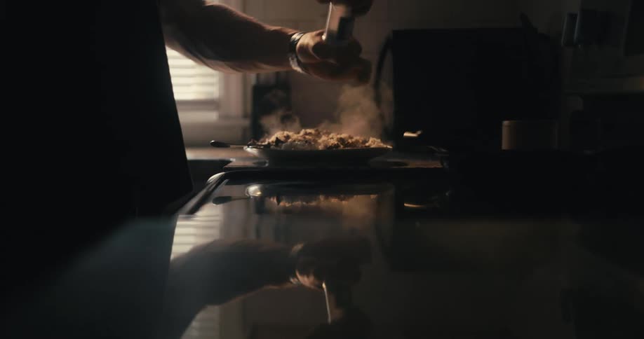 A man using a salt shaker on food he is preparing on a stove in the kitchen