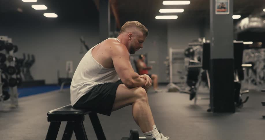 A man sitting on a black bench in a gym while lifting weights before putting them down on the floor