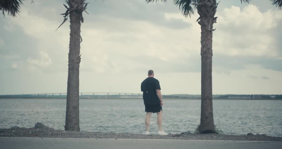 A man walking near water between two palm trees on a bright day with a cloud covered sky