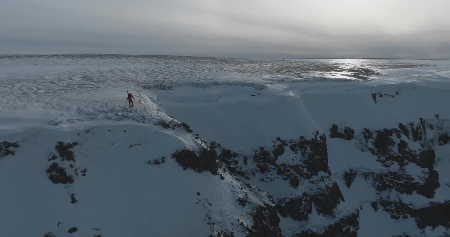 A man is walking through the snow with trekking poles to the edge of a cliff overlooking a river under a cloudy sky